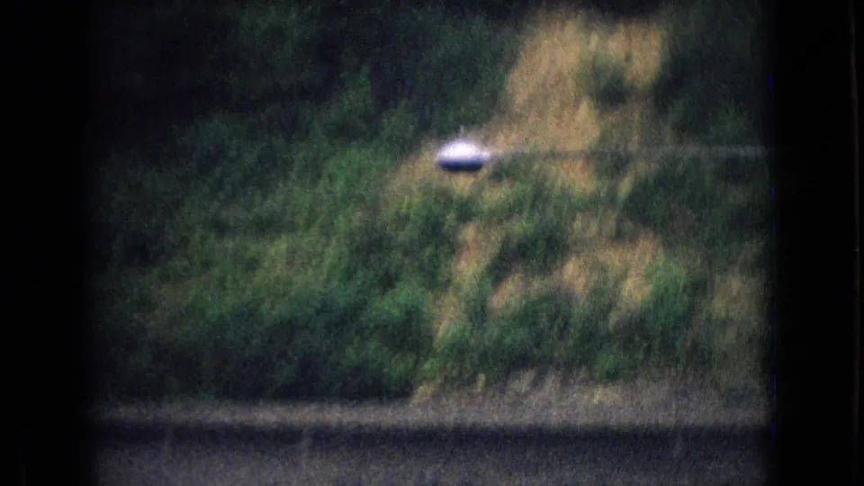 1965 New Jersey 8mm frame showing blurred roadside greenery, a dark foreground barrier, and a wooded hillside seen from a moving vehicle.