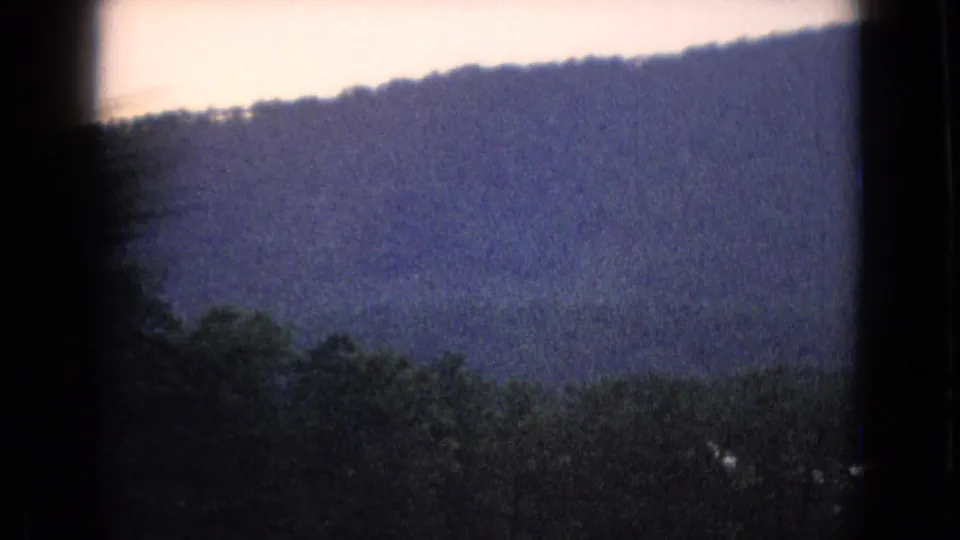 1965 New Jersey 8mm frame showing dark foreground trees, a distant wooded ridge, and pale sky filmed from a moving vehicle.
