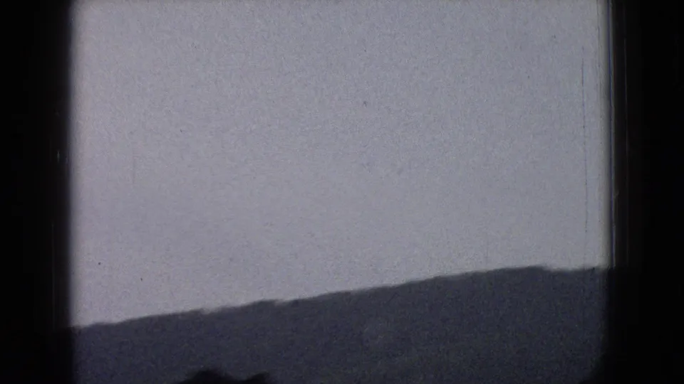 1965 New Jersey home-movie frame showing a silhouetted hillside or mountain ridge under a pale sky from a moving car perspective.
