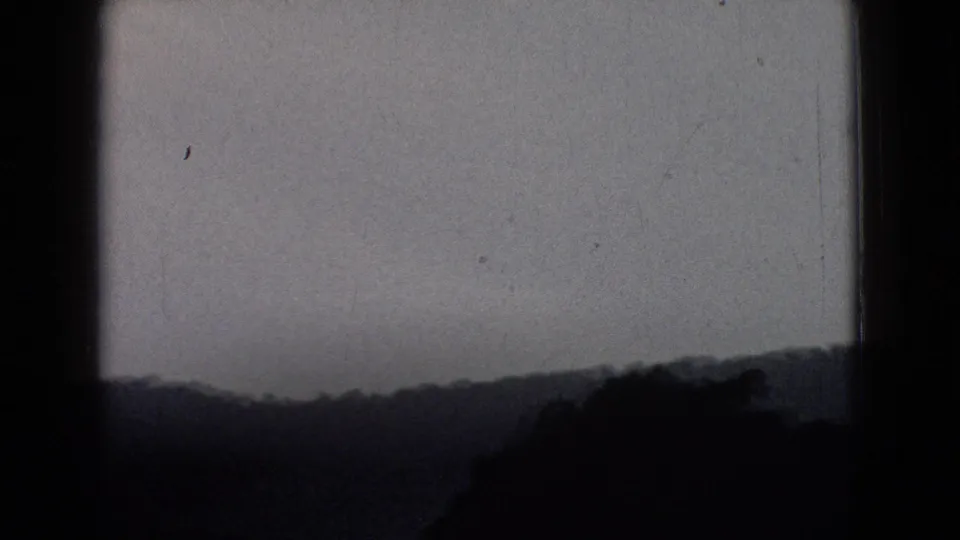 Grainy 1965 road-travel frame showing layered dark hills below a pale sky, likely filmed from a moving vehicle in New Jersey.