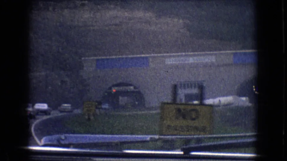 A moving 8mm frame from 1965 New Jersey showing a curving road, traffic, signs, a large roadside building, and a tunnel entrance ahead.