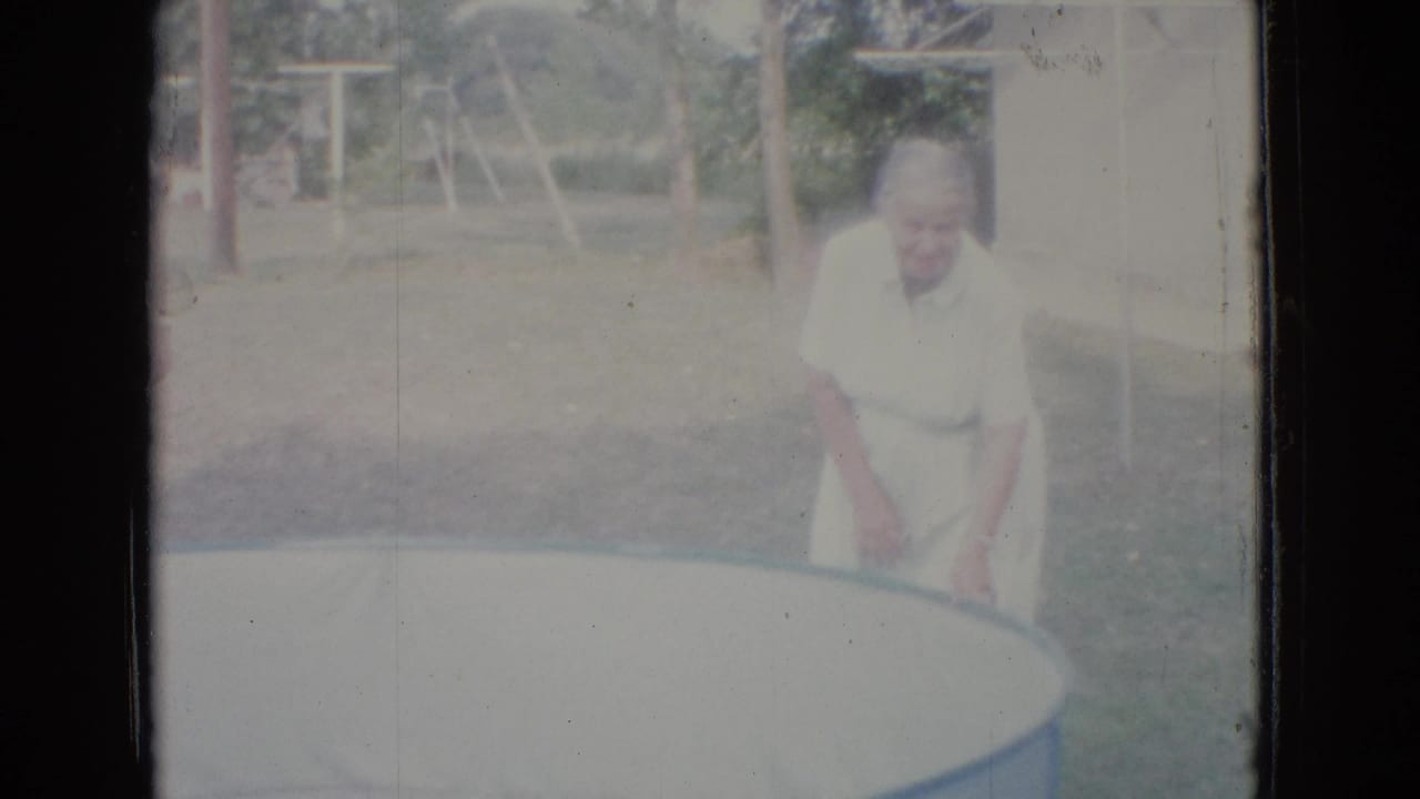 In 1965 New Jersey home-movie footage, an older woman in a light dress and apron stands near the edge of an empty above-ground pool in a grassy yard.