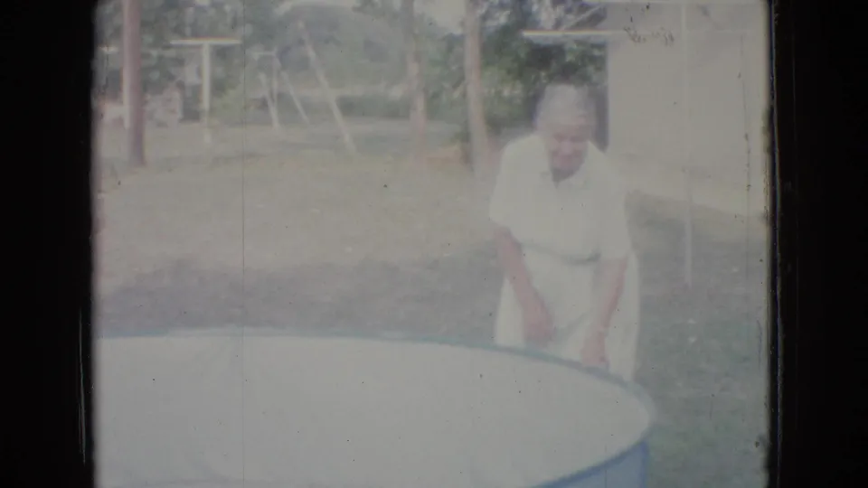 In 1965 New Jersey home-movie footage, an older woman in a light dress and apron stands near the edge of an empty above-ground pool in a grassy yard.