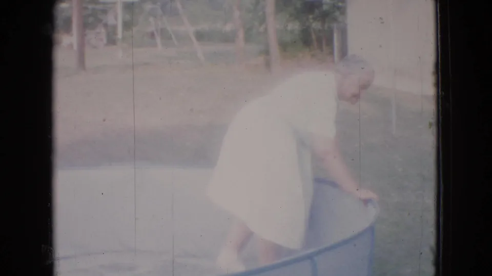 The older woman leans into the empty above-ground pool, appearing to adjust the liner or edge in a sunlit backyard setting.