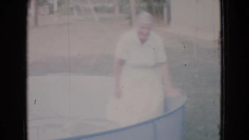 The woman stands close to the empty pool edge in the soft, faded 8mm frame, with grass and trees visible behind her.