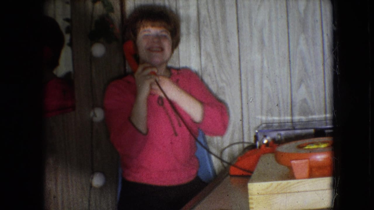 In this 1966 Athens, Ohio home-movie frame, a young woman with red hair stands near a red rotary phone and mirror while laughing in a wood-paneled interior.