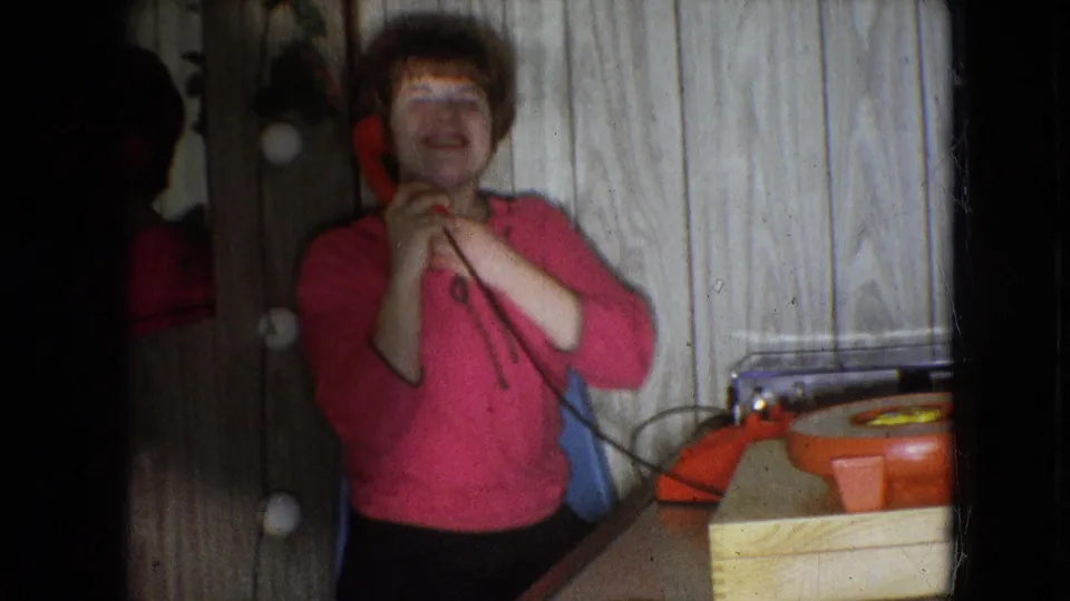 In this 1966 Athens, Ohio home-movie frame, a young woman with red hair stands near a red rotary phone and mirror while laughing in a wood-paneled interior.