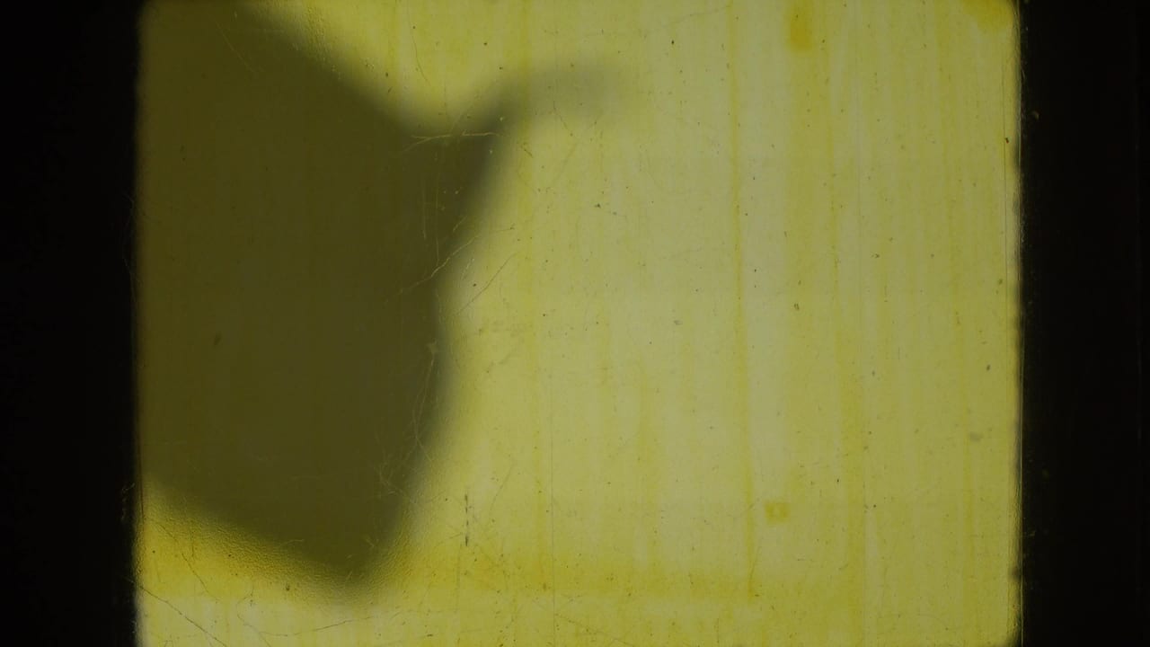 1966 Athens, Ohio 8mm frame showing a soft dark shadow across a yellow-tinted interior wall area with visible film grain and edge marks.