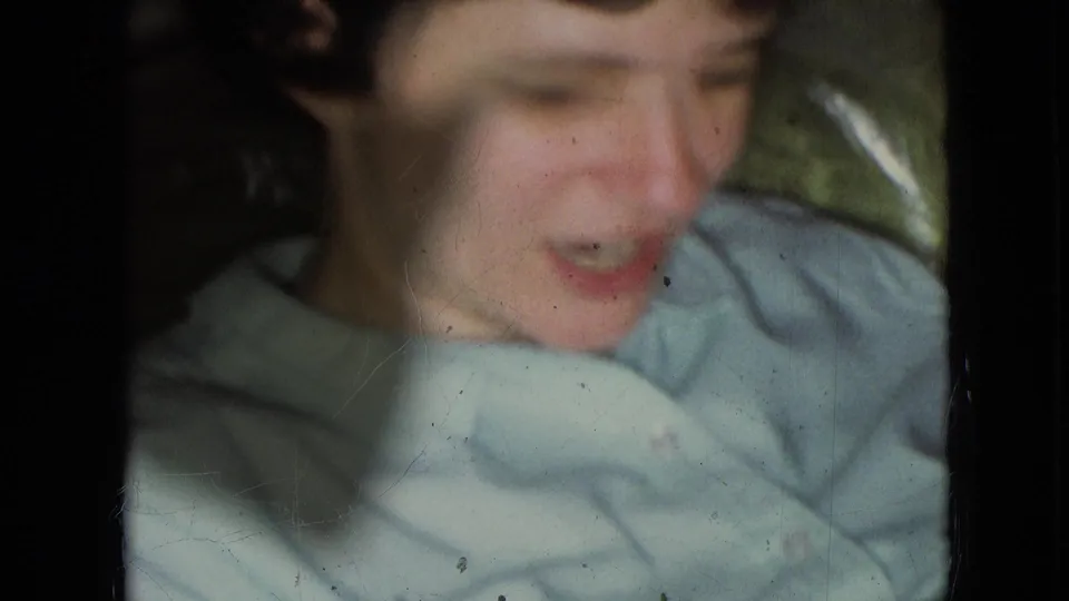 Close 8mm frame of the young boy in Athens, Ohio, appearing to speak while leaning near pale fabric, with scratches and grain visible.