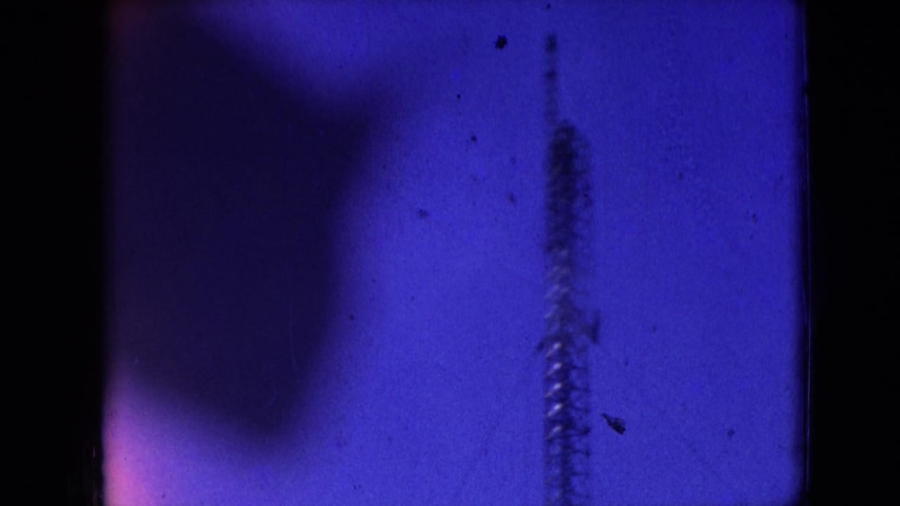 Athens, Ohio 1966 8mm frame showing a blue-tinted view of a faint vertical drilling structure or rig element behind dark foreground obstruction and film-edge shadows.