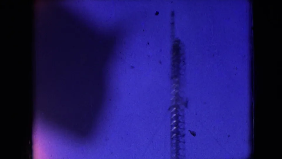 Athens, Ohio 1966 8mm frame showing a blue-tinted view of a faint vertical drilling structure or rig element behind dark foreground obstruction and film-edge shadows.