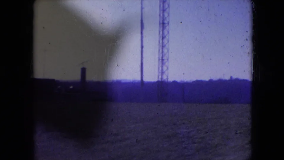 Athens, Ohio 1966 8mm frame showing an open field, distant treeline or low horizon, a tall drilling rig element, and dark foreground shadowing along the left edge.