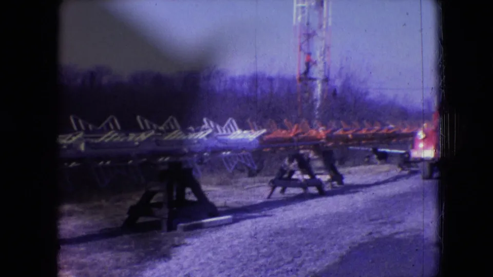 1966 Athens, Ohio frame with long metal framework on sawhorses, a tall lattice tower, a red-and-white truck edge, and open ground at a utility construction site.