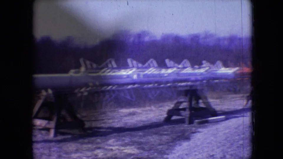 1966 Athens, Ohio home-movie frame showing metal framework sections arranged on wooden supports, with a distant tree line and small work-site details softened by the 8mm scan.
