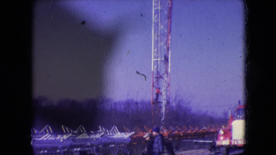 1966 Athens, Ohio 8mm frame showing a lattice communication tower, stacked metal sections, a distant worker, and a service truck with partial lettering.