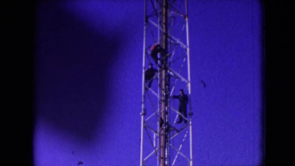 1966 Athens, Ohio 8mm frame showing several workers climbing or positioned on a metal lattice communication tower high above the ground.
