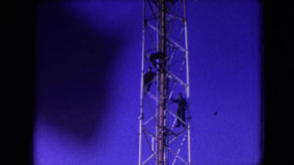 1966 Athens, Ohio 8mm frame showing three dark-clad men on a metal lattice tower, with one higher on the structure and two lower against a saturated blue sky.