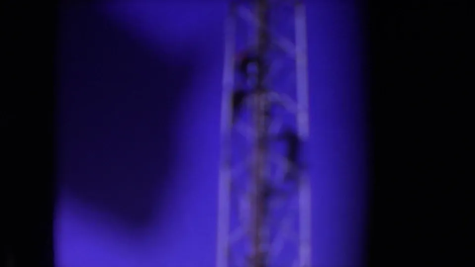 Soft 8mm frame from the Athens, Ohio clip showing a blurred metal tower segment and a dark figure climbing near the upper portion against a blue-purple sky.