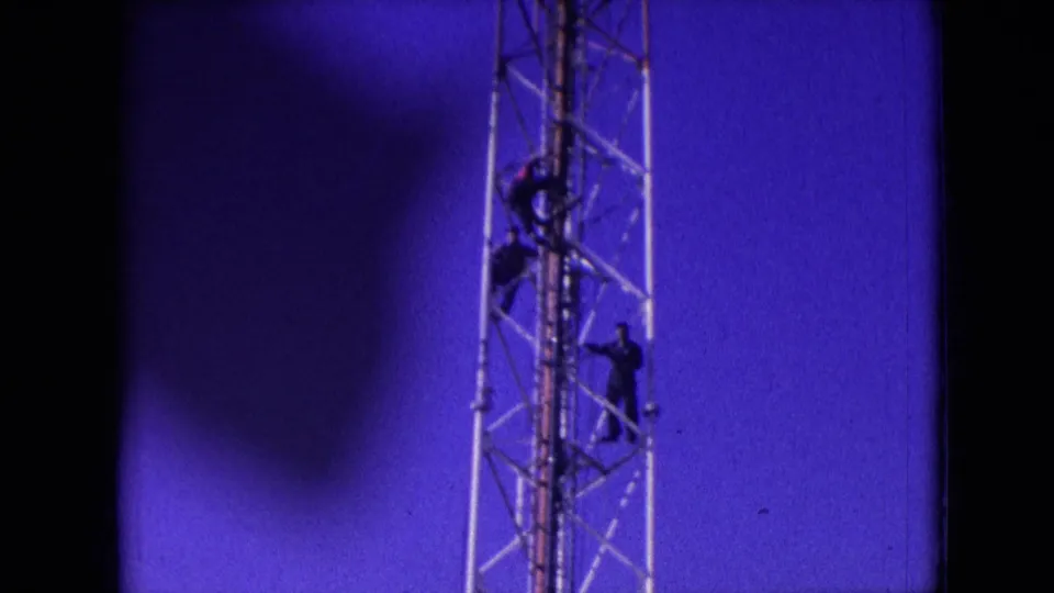 1966 Athens, Ohio 8mm frame showing three men spaced vertically on a metal lattice tower, with dark film borders and a vivid blue sky behind them.