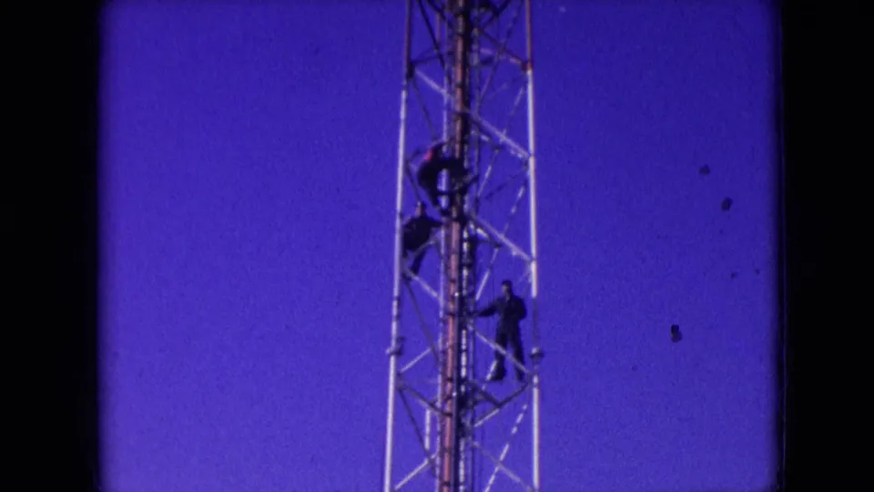 Final 1966 Athens, Ohio 8mm frame showing three dark-clad men climbing or holding positions on a metal lattice tower, with dust marks and grain visible over the blue sky.