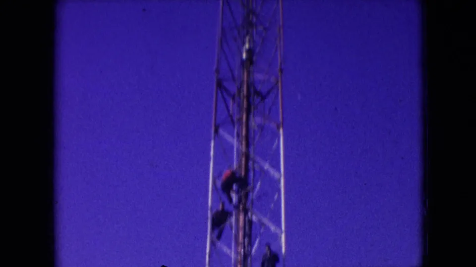 Athens, Ohio 1966 home-movie frame showing a worker on a tall lattice radio tower against a saturated blue-purple sky with visible 8mm film borders.