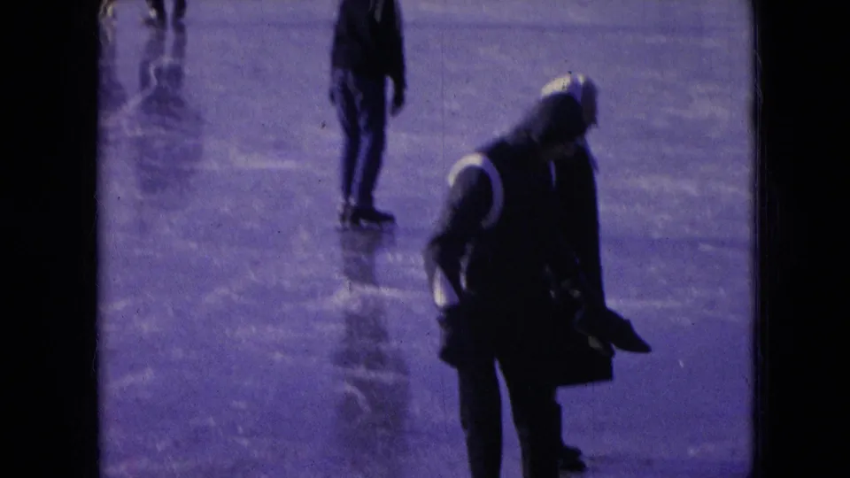 A bundled skater bends forward on the frozen surface while another figure stands behind, captured in soft purple-tinted 8mm footage from Athens, Ohio in 1966.