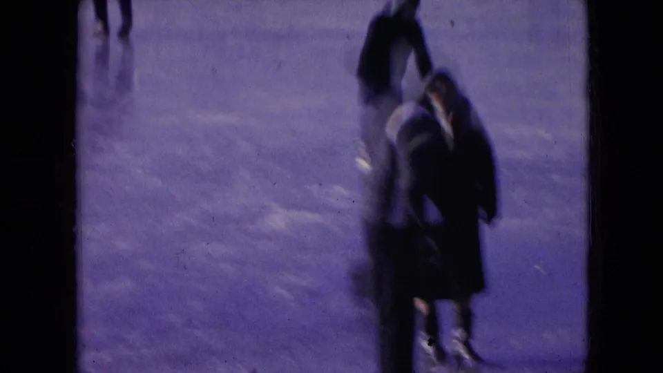 Two bundled skaters appear close together on the ice, with motion blur, dark coats, and the purple-blue cast of 1966 Athens, Ohio 8mm home-movie film.