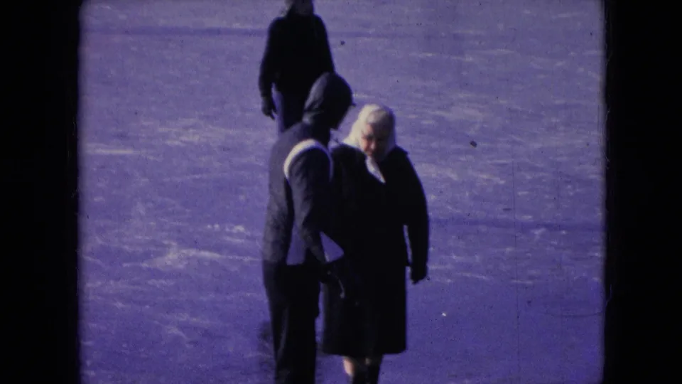 In supplied 1966 Athens, Ohio 8mm footage, a hooded man and a woman in a white headscarf walk together across a frozen snow-covered surface while another figure passes behind them.