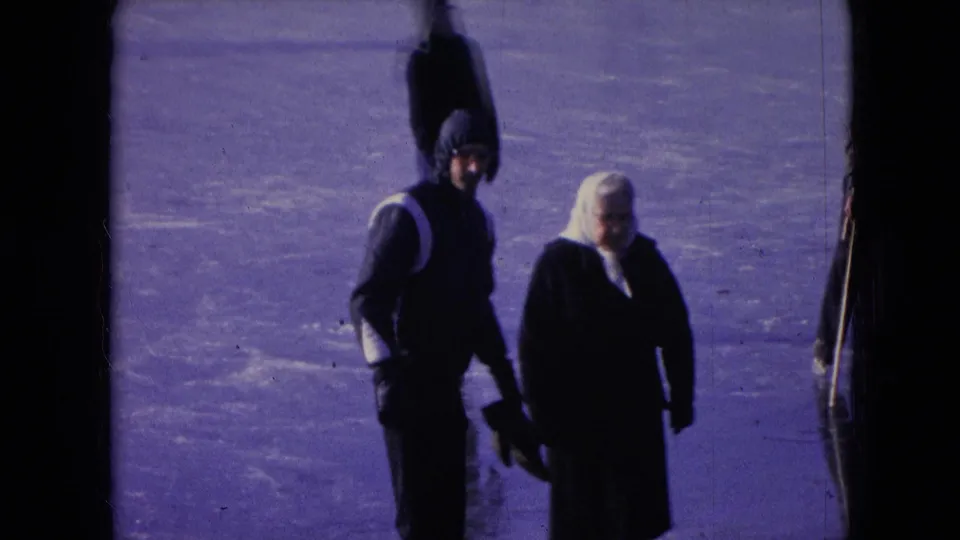In the Athens, Ohio home-movie frame, a man in a hooded jacket and a woman with a white headscarf move across the frozen surface with a distant figure behind them.