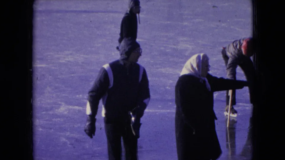 The 1966 home-movie frame shows a hooded man with gloves and a woman in a white headscarf on a frozen surface, with the woman reaching toward a vertical edge or support at the right.