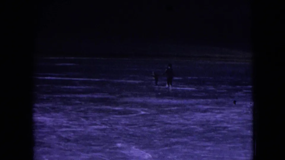 A wide, underexposed 8mm frame shows small distant figures moving across a frozen surface, with heavy shadow, purple-blue tint and black film borders dominating the view.