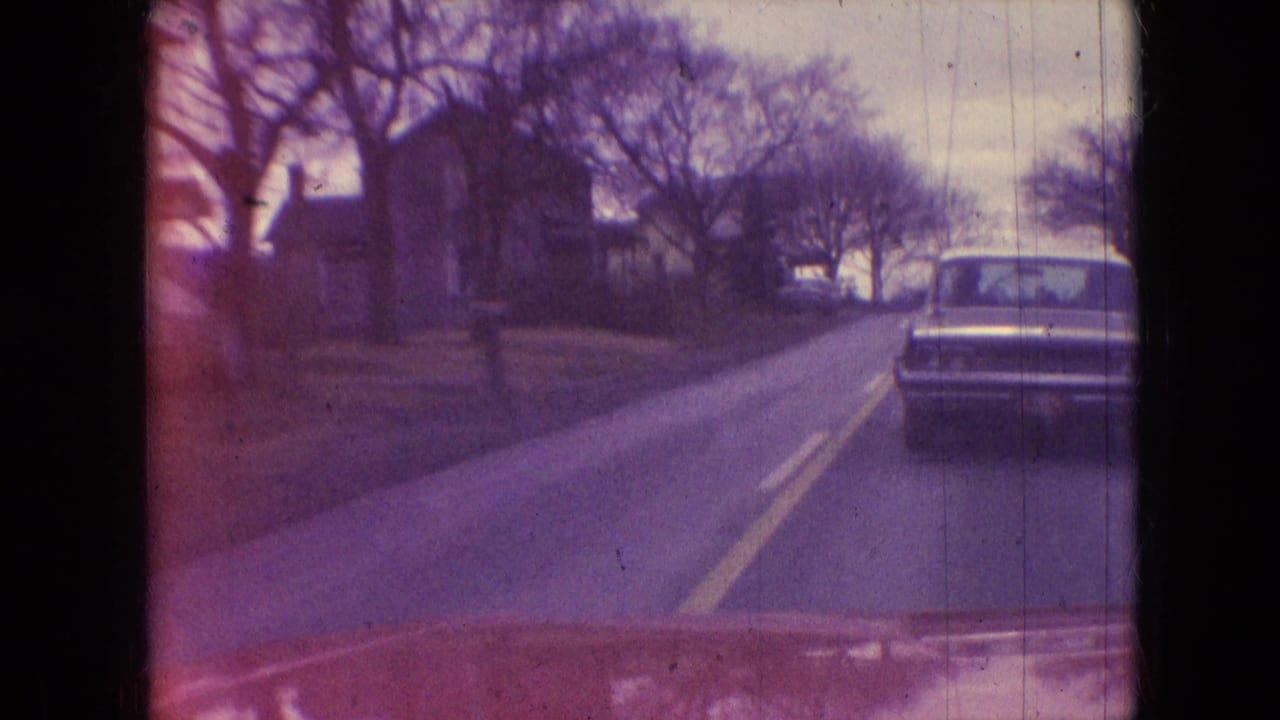 1966 Athens, Ohio home-movie frame looking through a windshield at a sedan ahead on a quiet residential road with bare trees and houses.