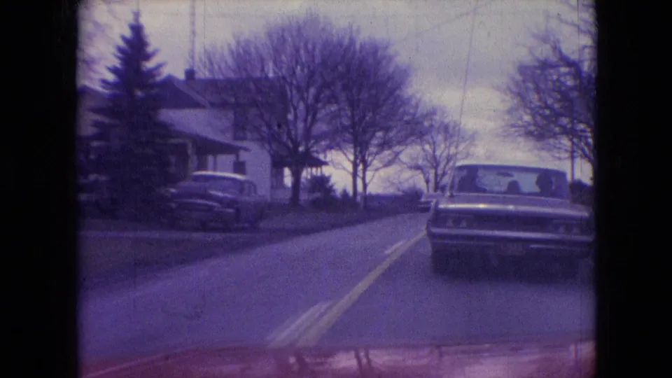 1966 Athens, Ohio street view from a moving car, with a sedan ahead, houses to the left, bare trees, and soft purple 8mm color.