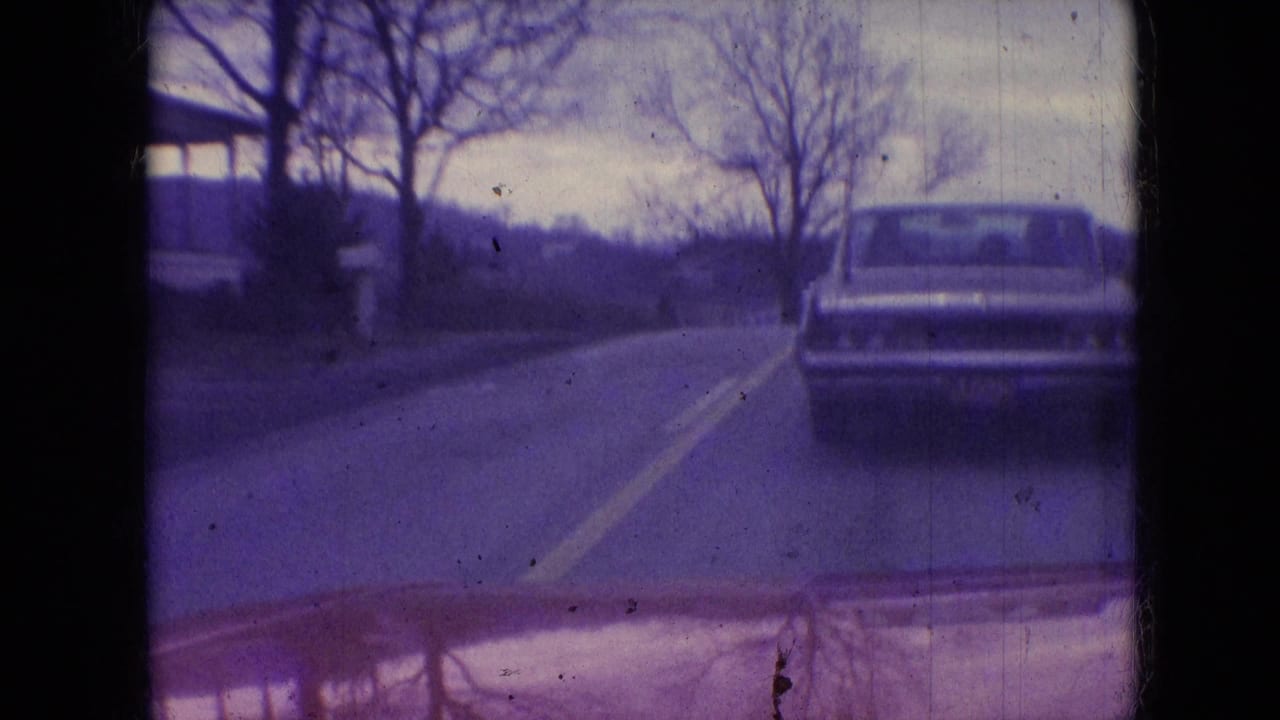 1966 Athens, Ohio home-movie frame from a moving car showing a classic sedan ahead on a two-lane rural road bordered by bare trees and distant hills.