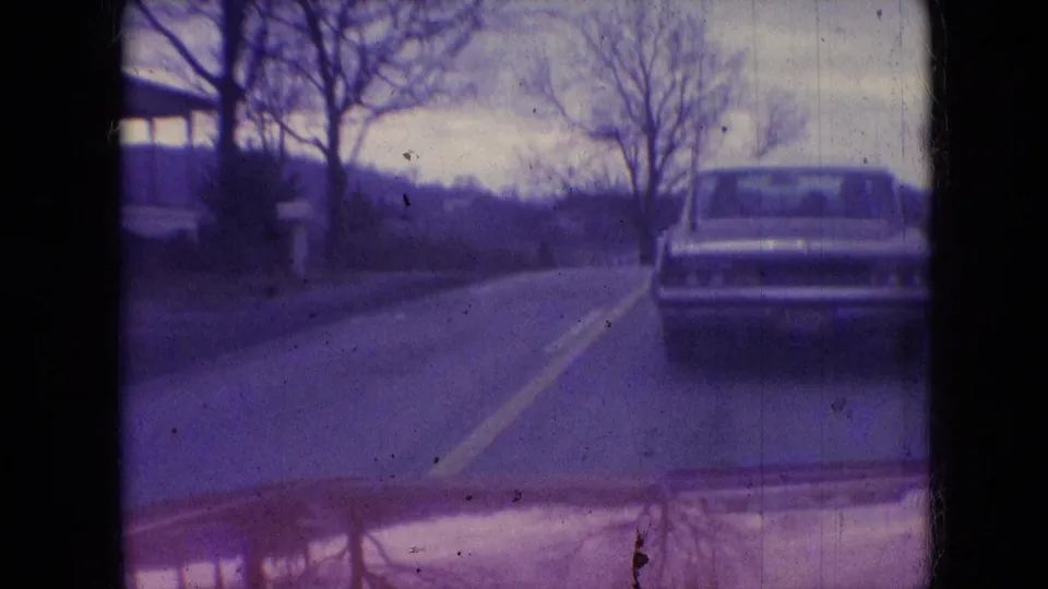 1966 Athens, Ohio home-movie frame from a moving car showing a classic sedan ahead on a two-lane rural road bordered by bare trees and distant hills.