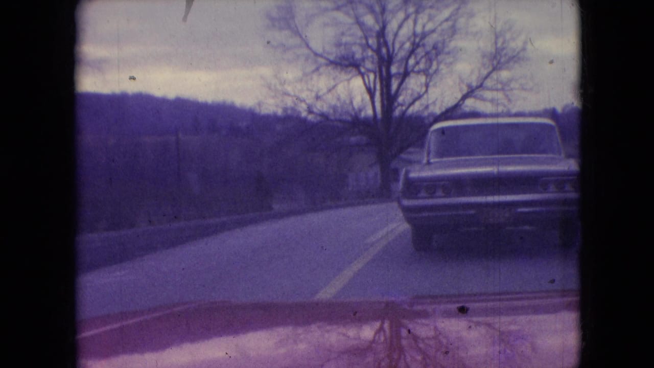 1966 Athens, Ohio frame showing a classic sedan traveling ahead on a gently curving rural road, with leafless trees and open land visible through aged 8mm color.