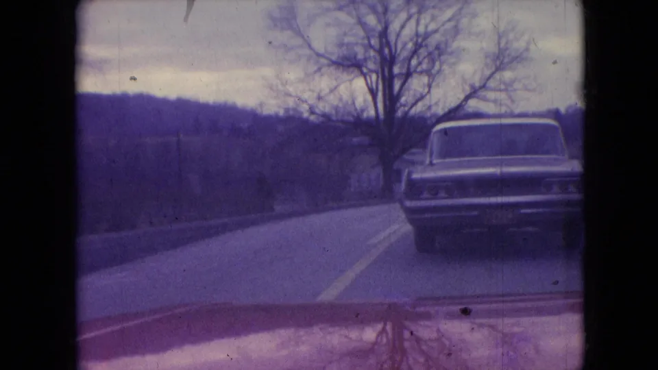 1966 Athens, Ohio frame showing a classic sedan traveling ahead on a gently curving rural road, with leafless trees and open land visible through aged 8mm color.