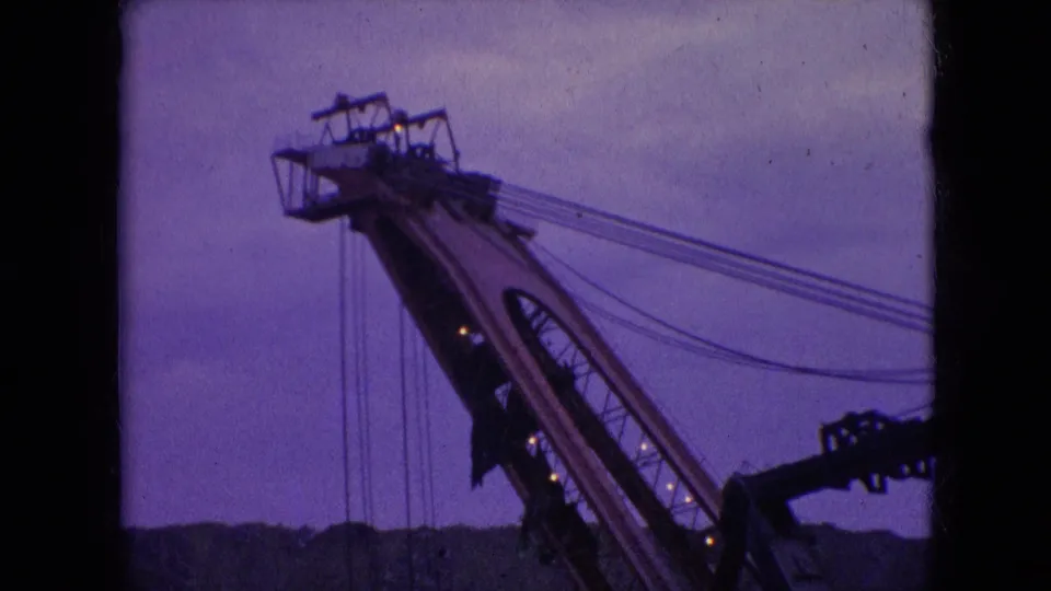 1966 Athens, Ohio 8mm frame of a tall construction crane upper section, with parallel cable lines stretching across a purple-blue evening sky.
