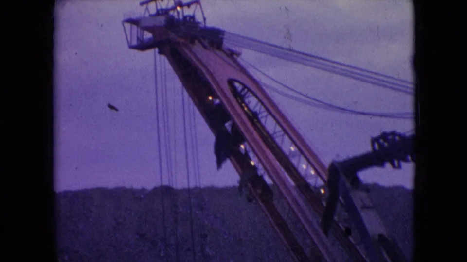 1966 Athens, Ohio frame with the excavator boom lifted above a dark quarry ridge, showing cables, machinery structure, and soft purple film color.