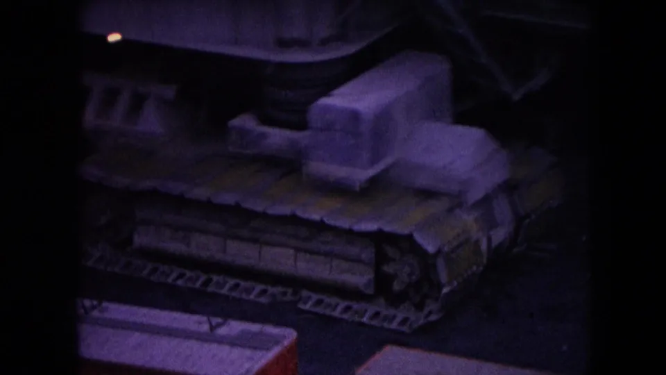 1966 Athens, Ohio frame focused on the excavator's crawler tracks, lower body, boom base, and bucket linkage in dark blue-purple 8mm color.