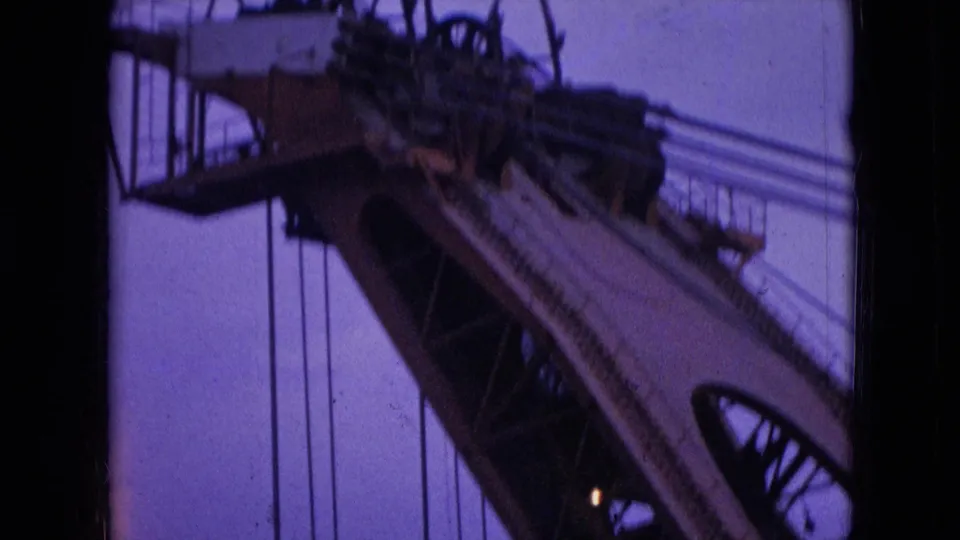 1966 Athens, Ohio 8mm frame showing a soft-focus close view of crane machinery, cable runs, a small platform, and angled steel members against the dusk sky.