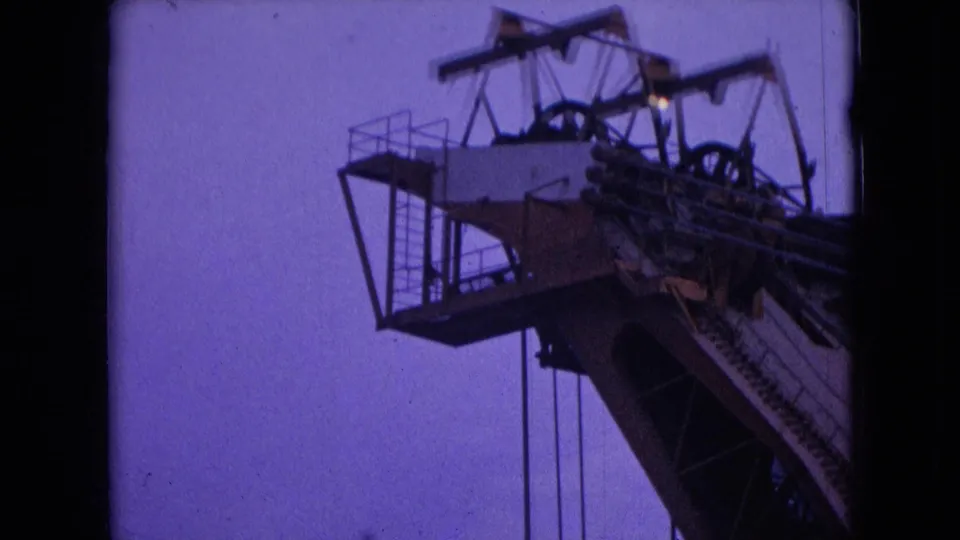 1966 Athens, Ohio 8mm frame showing the upper portion of a crane with a work platform, vertical cables, large pulley assembly, and steel boom against a violet dusk sky.