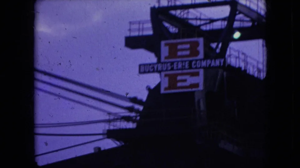 1966 Athens, Ohio 8mm frame showing a Bucyrus-Erie Company sign on a dark industrial machine structure with railings, wires, and a small greenish light visible nearby.