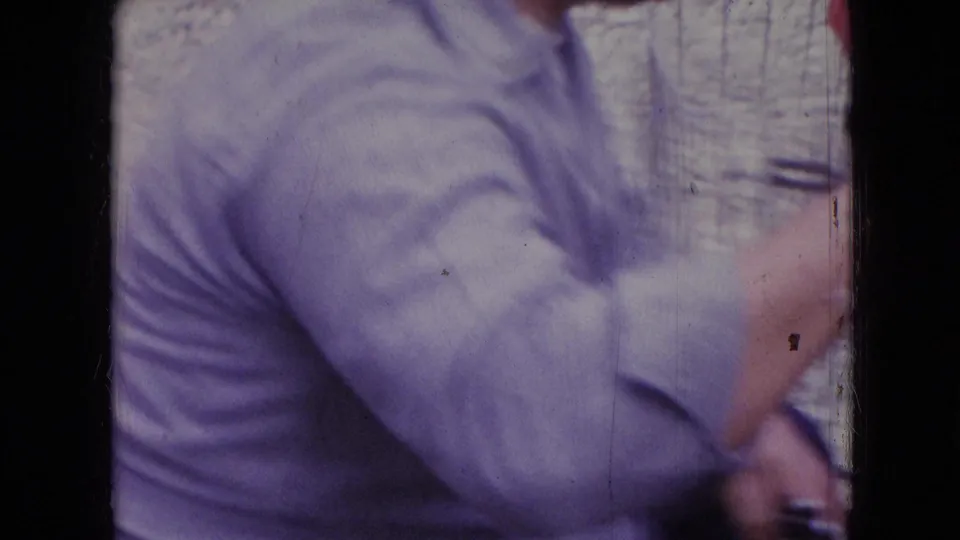 The man in a light purple shirt bends or kneels close to the car, his face out of frame, as the handheld 8mm image holds on his torso and arm.