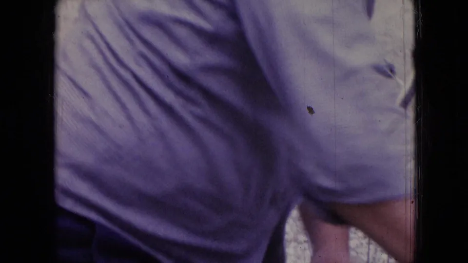 A close, partial 8mm frame shows the man’s torso and arm in a light purple shirt near the car, with soft focus and visible film borders.