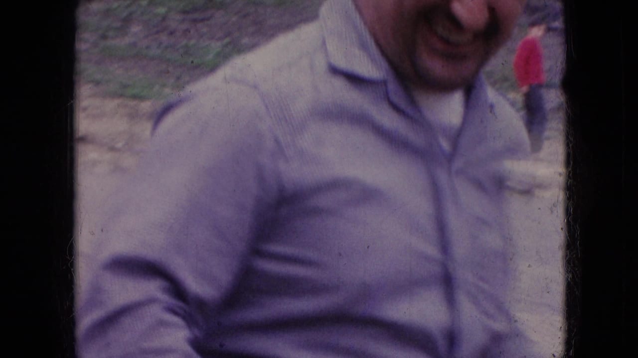 Man in a light shirt leaning near a bicycle outdoors with another person in red in the background.