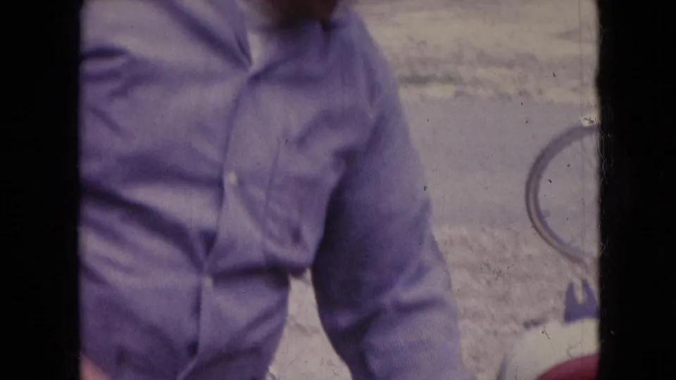 A close handheld frame shows the man in a light shirt beside a bicycle wheel on a pale roadside or gravel surface in the 1966 Athens, Ohio footage.