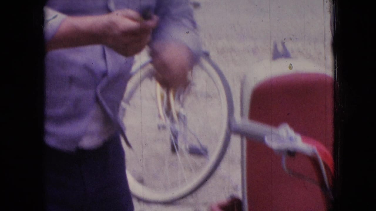 In supplied 1966 Athens, Ohio 8mm footage, a man in a light blue shirt is partly framed while working near a bicycle and a red scooter on a residential roadside.