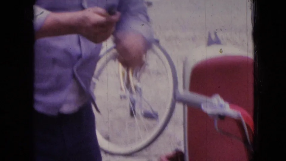In supplied 1966 Athens, Ohio 8mm footage, a man in a light blue shirt is partly framed while working near a bicycle and a red scooter on a residential roadside.