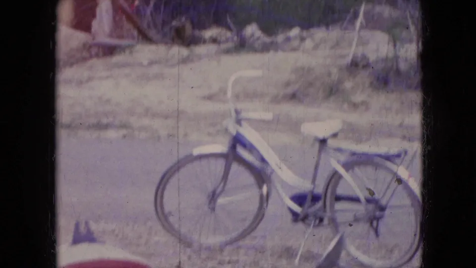 The white bicycle with a red saddle is seen more fully on the roadside in the supplied 1966 Athens, Ohio home-movie footage, with part of the red scooter entering the lower frame.